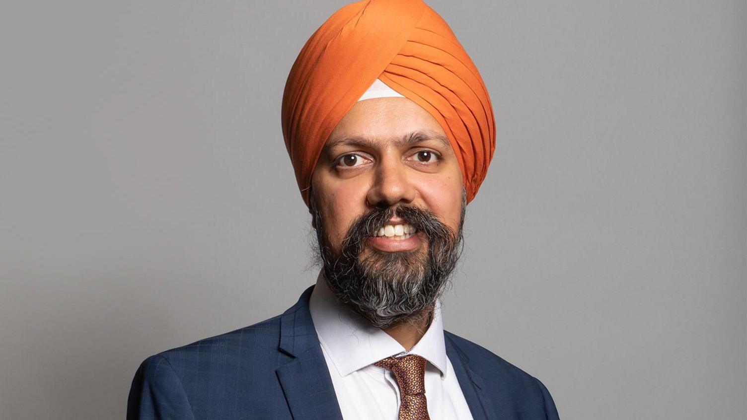 A headshot of a man with a greying beard wearing a blue suit and an orange turban.