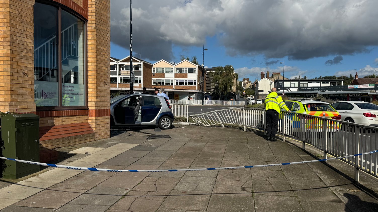 A navy Smart car smashed into a brick building while a police officer looks out into the road. Blue and white police tape cordons off the scene. 