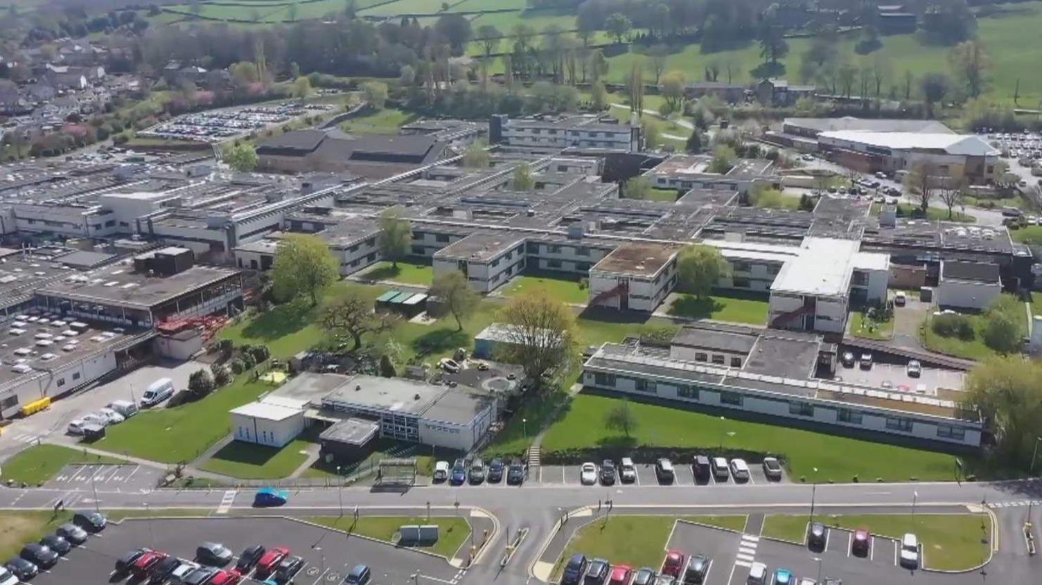 An aerial view of lots of connected low rise buildings at the Airedale Hospital site. Cars parked in areas outside most of the buildings. Trees and some areas of grass between some of the buildings.