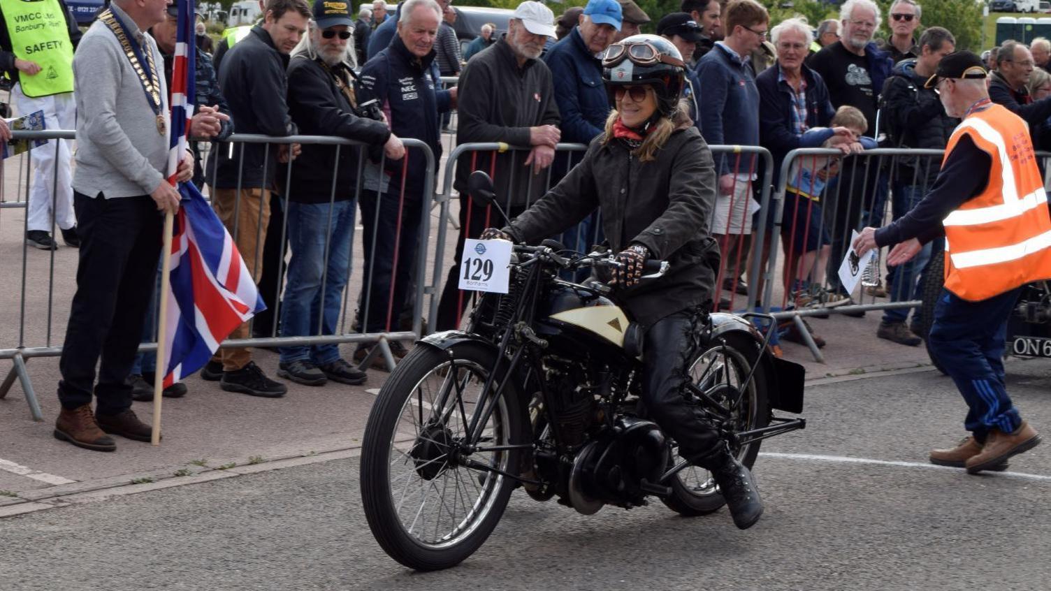 A woman wearing a black jacket, what look like black leather trousers and black boots sits on a vintage motorcycle. She is wearing a helmet with a pair of goggles perched on it. Crowds of people are watching from the other side of a barrier.