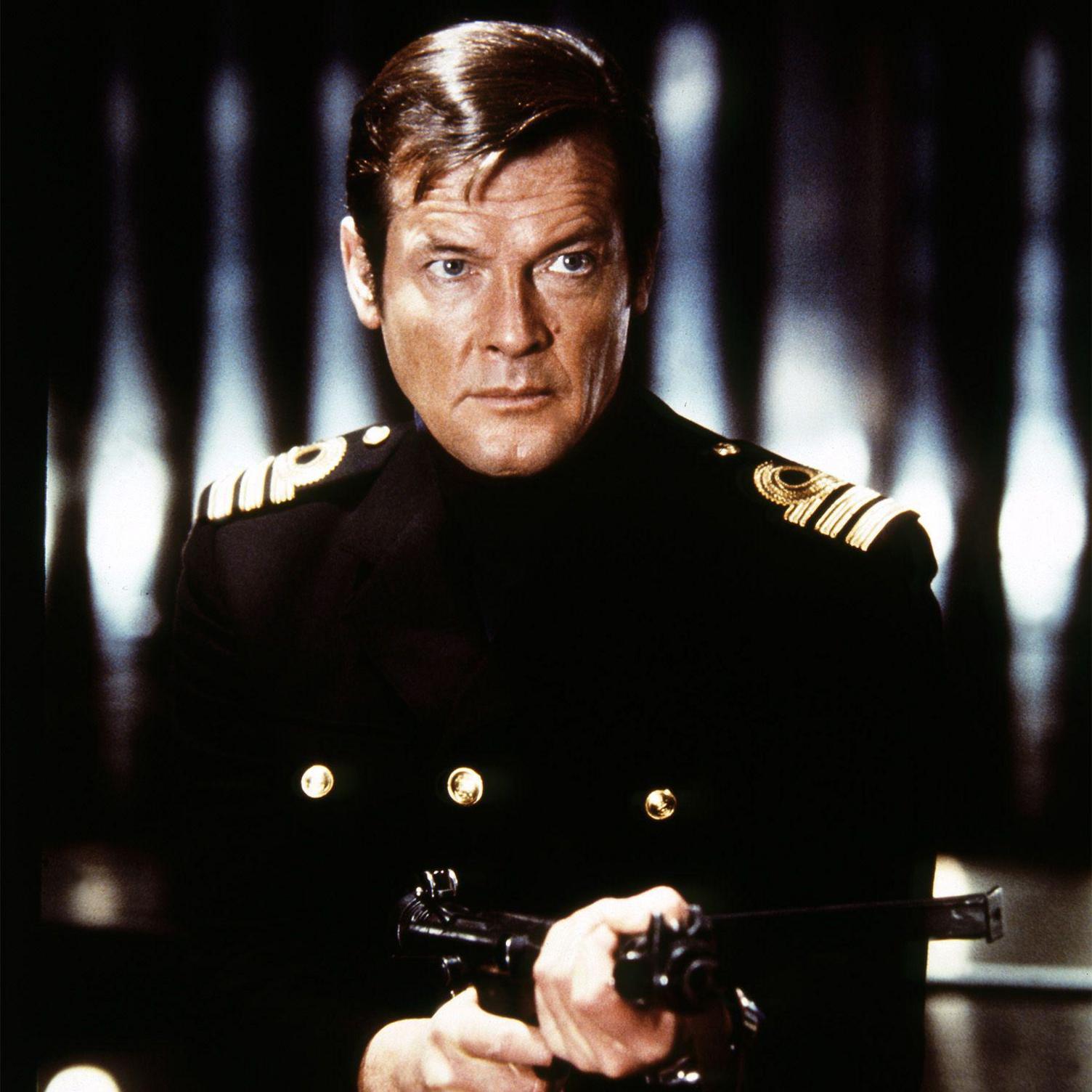 Sir Roger Moore is seen on the set of the James Bond film, The Spy Who Loved Me. He is wearing a double-breasted naval officer's uniform and is pointing a gun at someone off camera. He has a serious and purposeful expression on his face. 