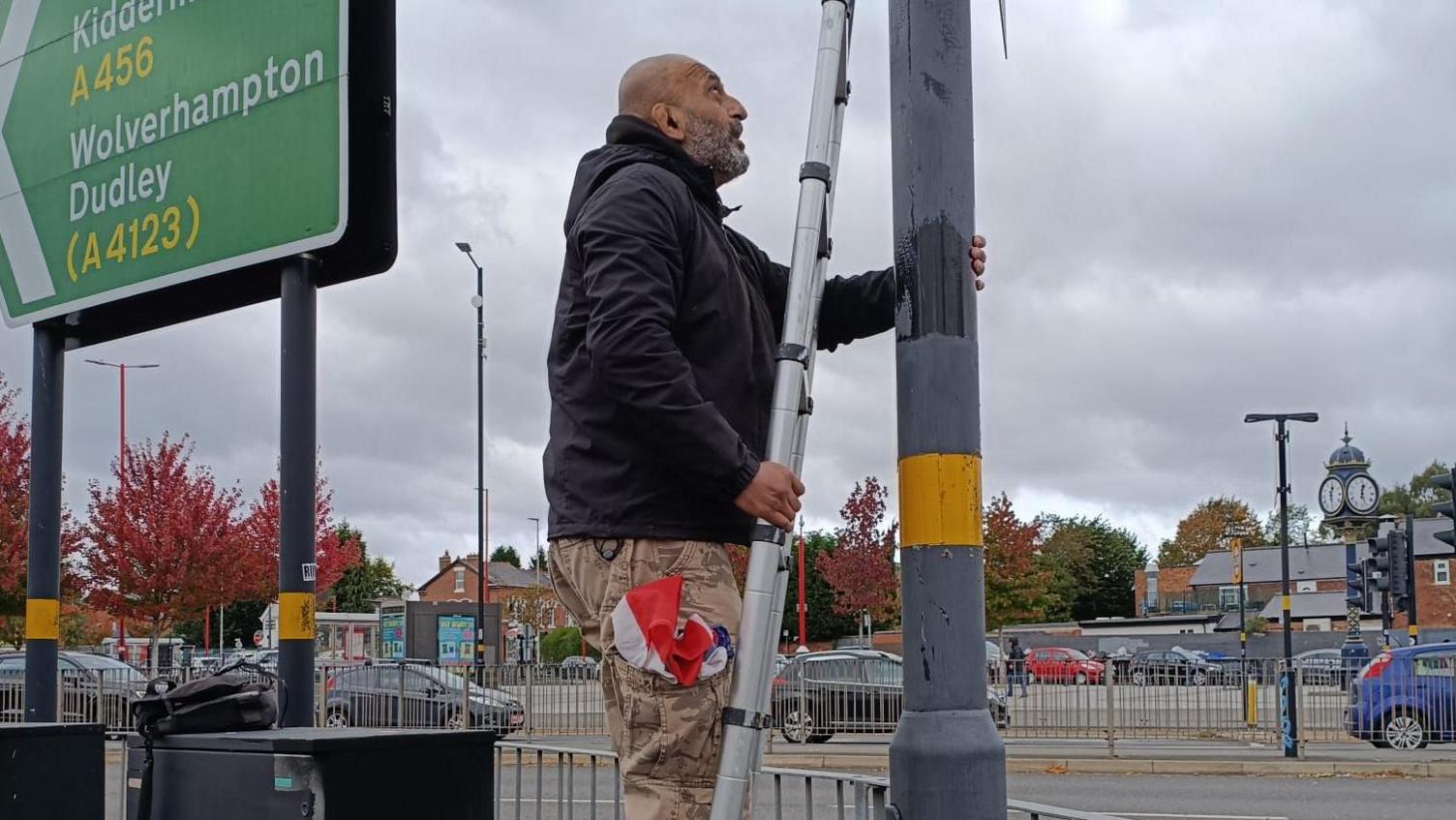 Salman Mirza on a ladder taking flags down from a lamp-post. He has what looks like a union jack flag in the side pocket of some yellow cargo trousers. We can see he is on a busy residential road. There is a sign next to him giving directions to Kidderminster, Wolverhampton and Dudley.