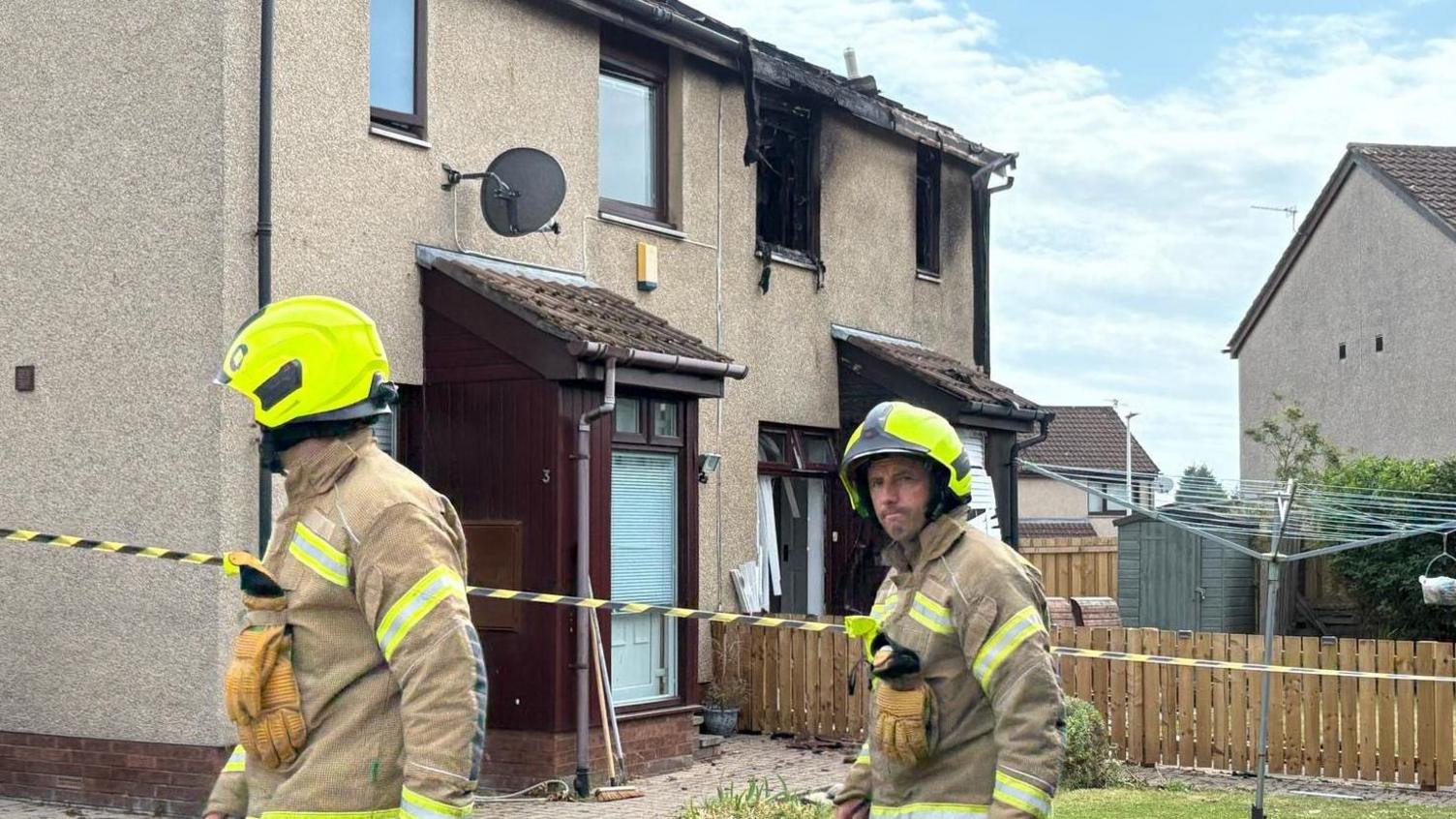 Two firefighters walk past a taped off house. The top two windows have been smashed and fire-damaged