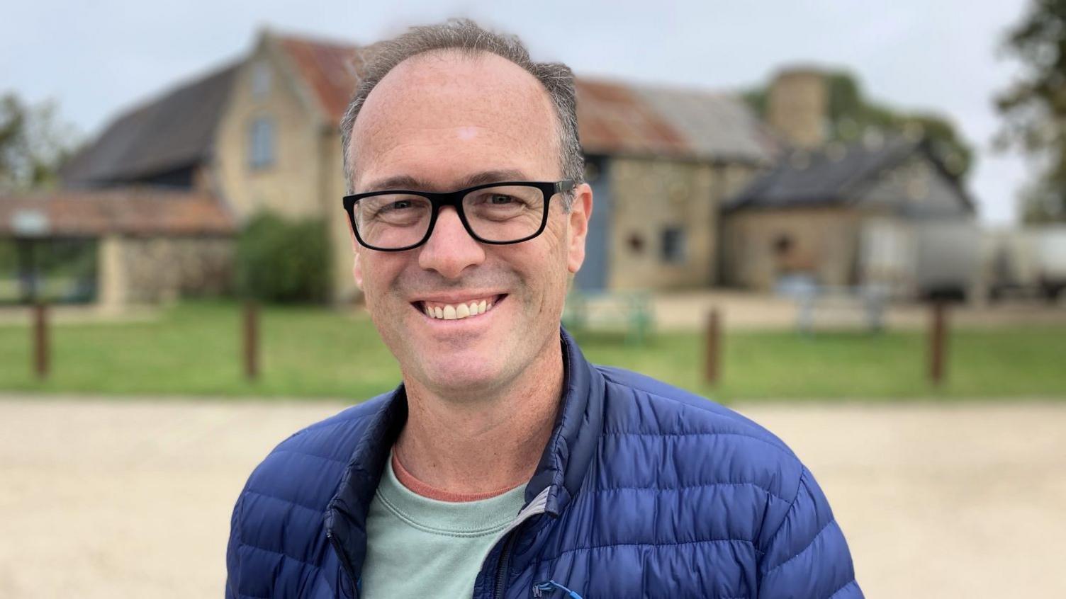 A head and shoulders photo Hektor Rous. He has short greying hear and black glasses. He is wearing a blue padded jacket and a light green top underneath, and he is stood in front of an old farm building