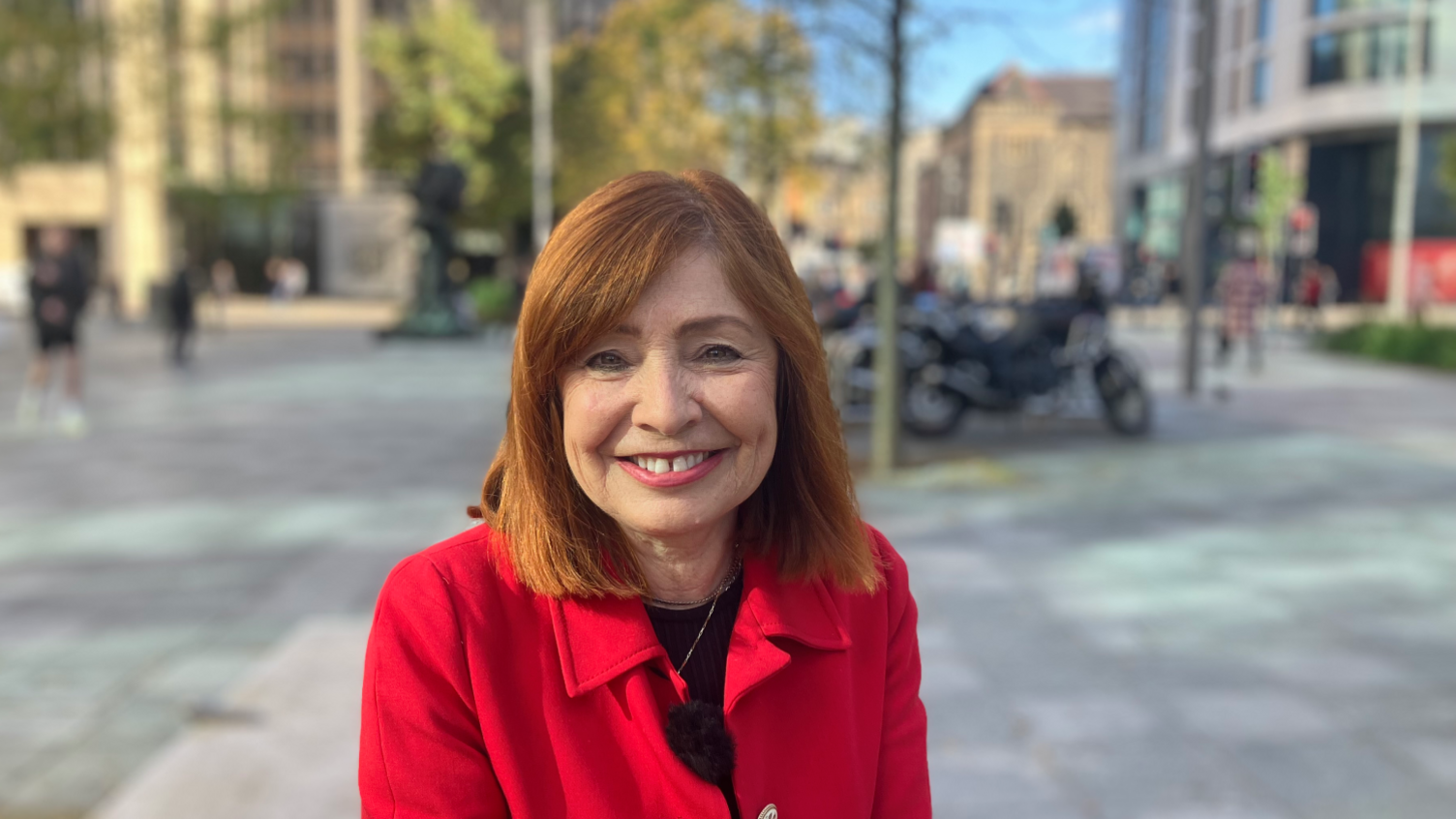 Sandra Busby of Cnect Wales, wearing a red coat and smiling at the camera in the street, surrounded by office buildings