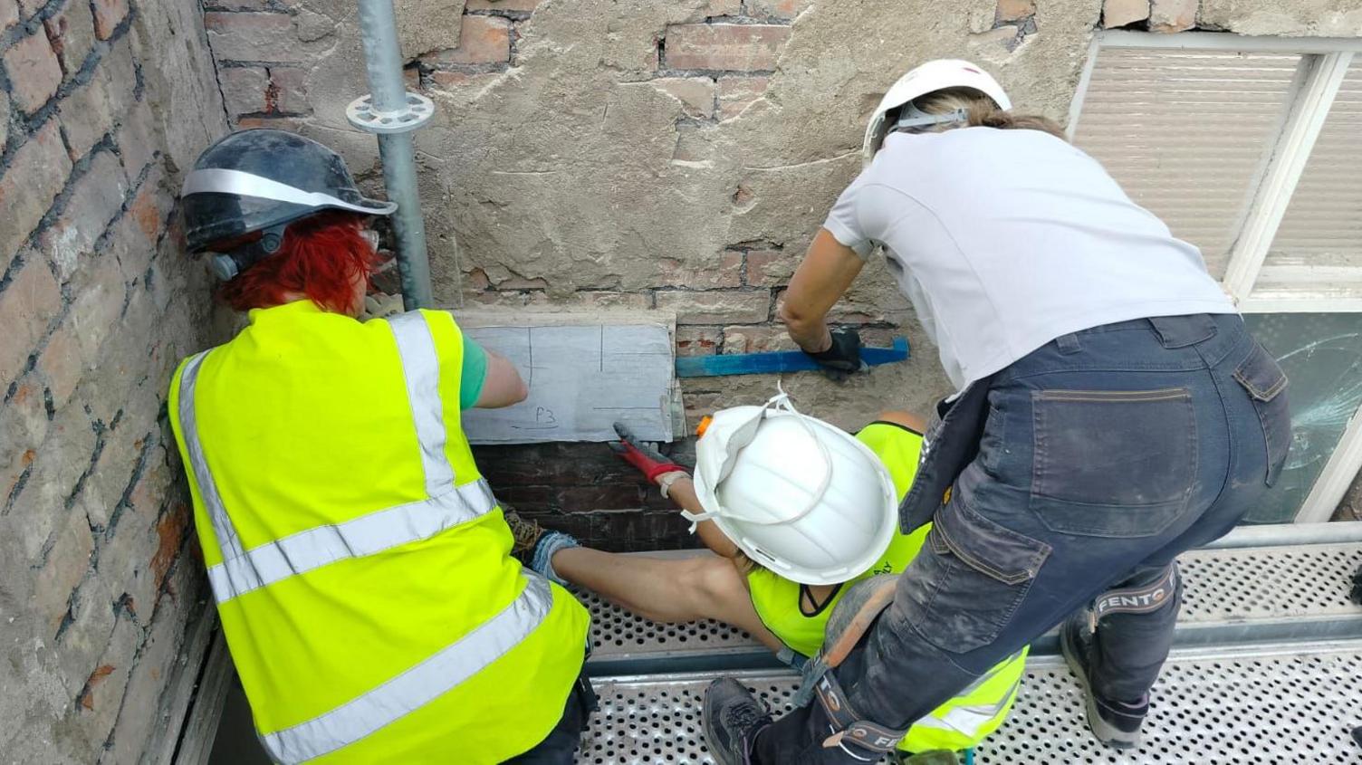 Three people stand on decking as they hold a piece of the mosaic as they prise it carefully from a brick wall. They are wearing hard hats and high-vis vests.