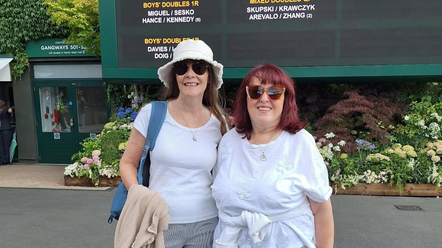 Sherly and Marianne are standing in front of a fixtures board in the Wimbledon grounds