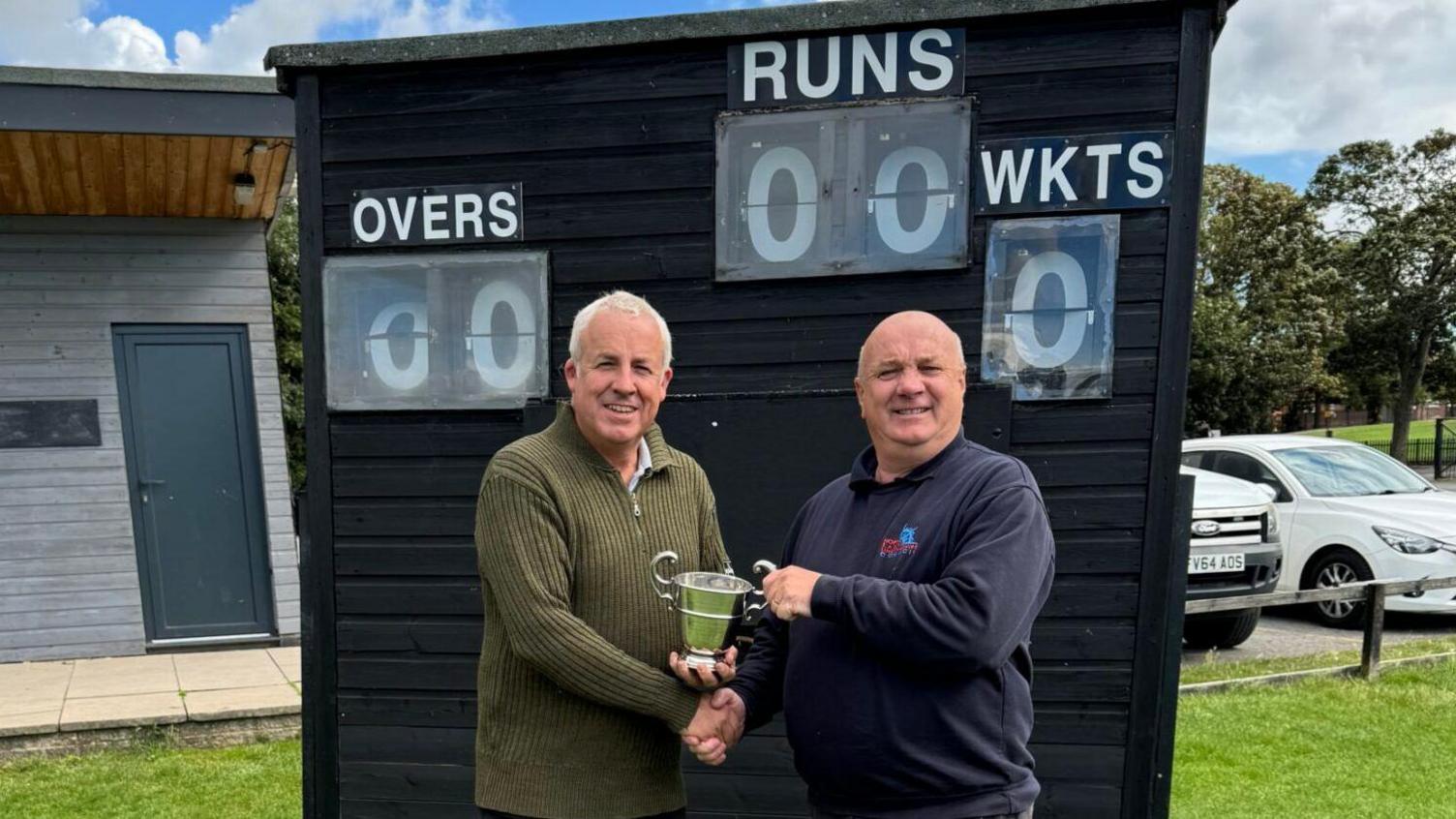 The two men shake their right hands whilst exchanging the cup with the other.  They smile at the camera in front of a cricket score board with nil overs, runs or wkts displayed.