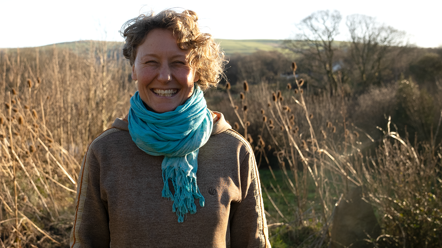 Rosie is standing in the grounds of Brithdir Mawr surrounded by grass and bushes with hillside behind her.  She has short curly fair hair and is wearing a brown long-sleeved top and a blue cotton scarf. 