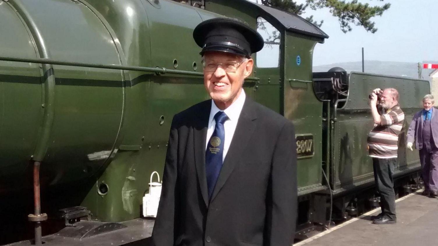 Mr Bartlett is standing in front of a green steam train. He is wearing a black suit and a conductor hat.