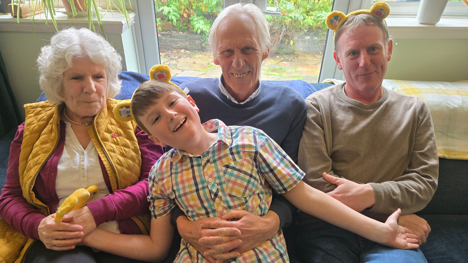 Taylor sitting in between his dad and family members. He is wearing yellow Pudsey ears and they are all sat on a sofa looking at the camera smiling. 