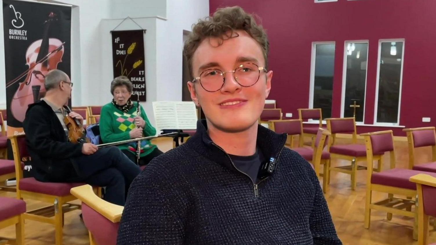 Man wearing glasses sitting in rehearsal room with two musicians behind him and several rows of empty chairs