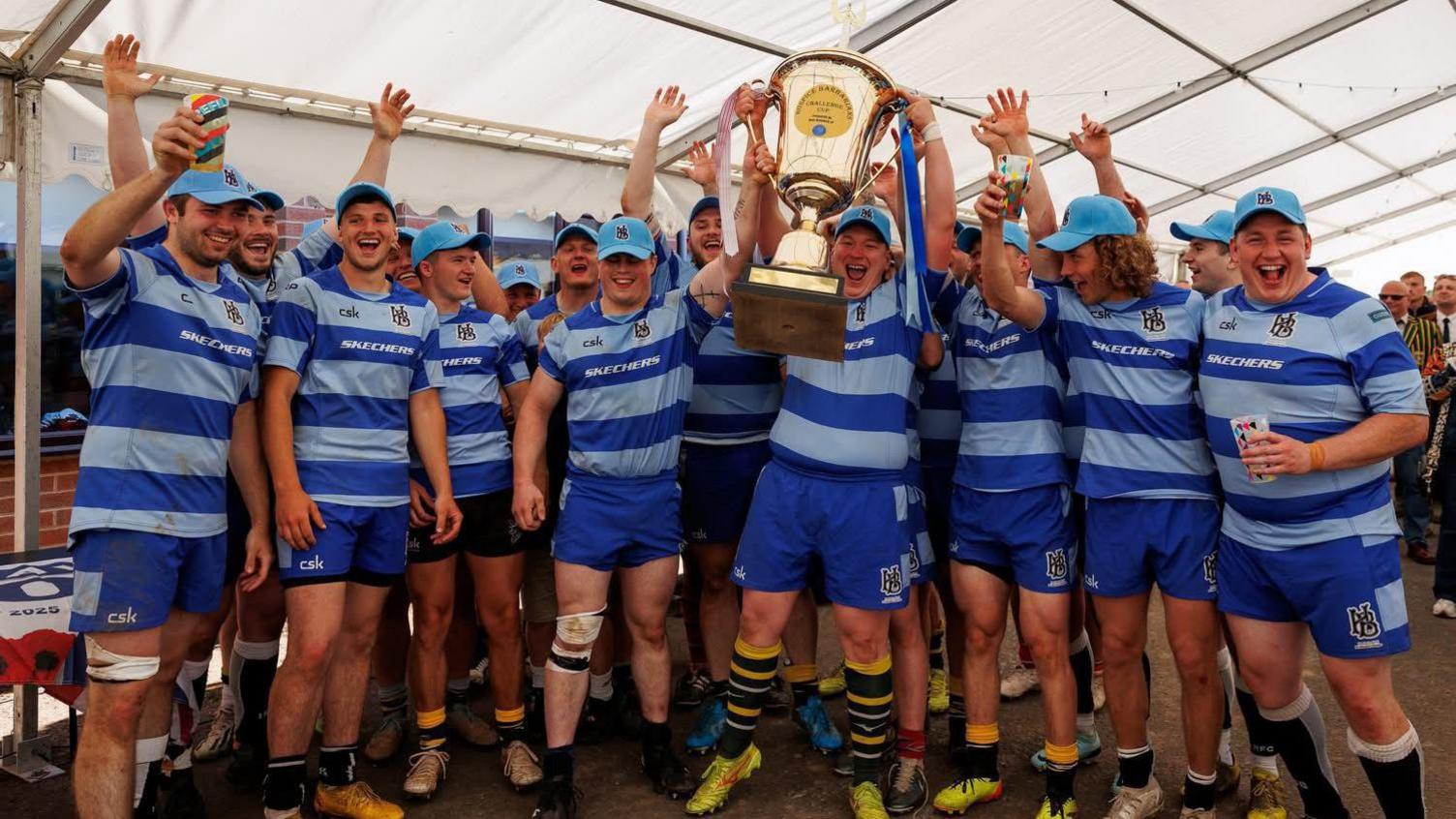 A team photograph of Hospice Barbarians, who wear a striped blue kit with the logo on the top. The team are celebrating together and holding a cup in a marquee.