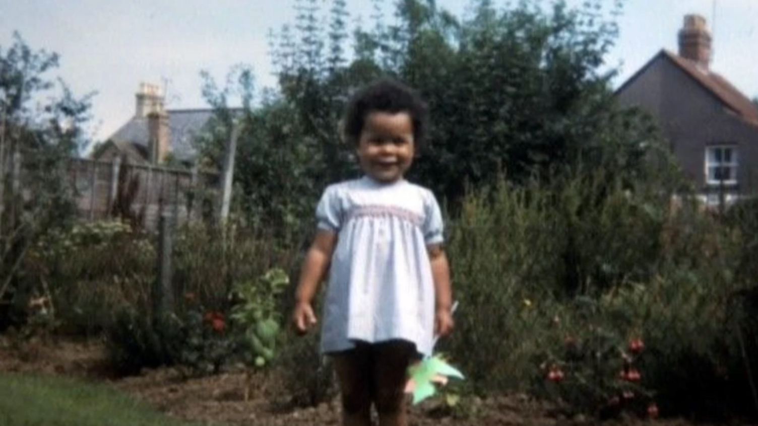 Vik Fielder standing in a garden as a young girl, wearing a dress and smiling. She has short black hair and is holding a windmill toy.