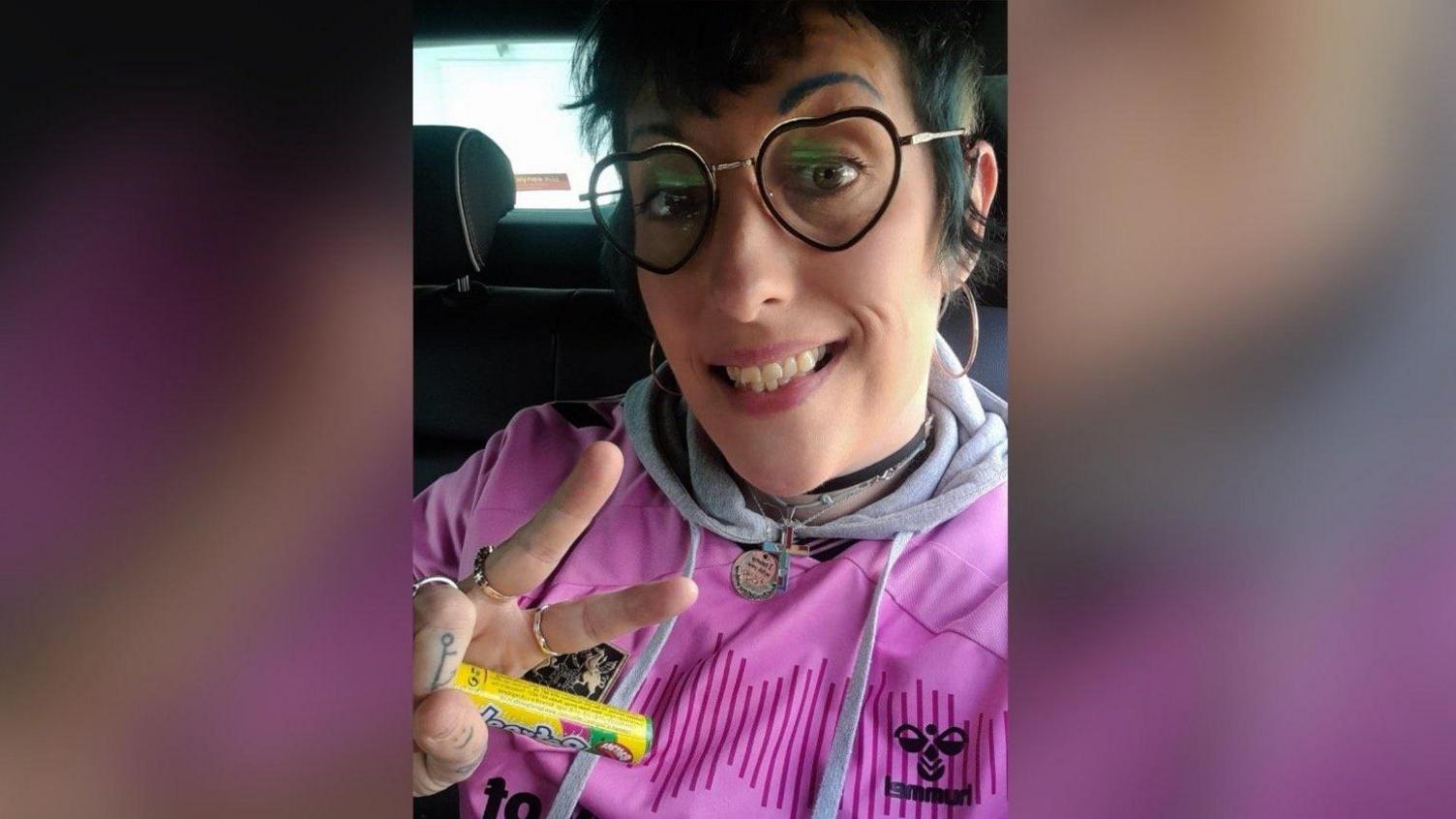 Heidi Green in a pink Northampton Town shirt and heart-shaped glasses. She is making a "peace" sign.