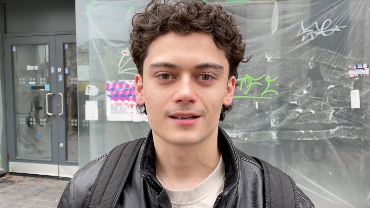 Joe Savory. A man with brown curly hair who is looking at the camera and smiling. He is wearing a black leather jacket and is wearing a backpack. He is stood outside. 