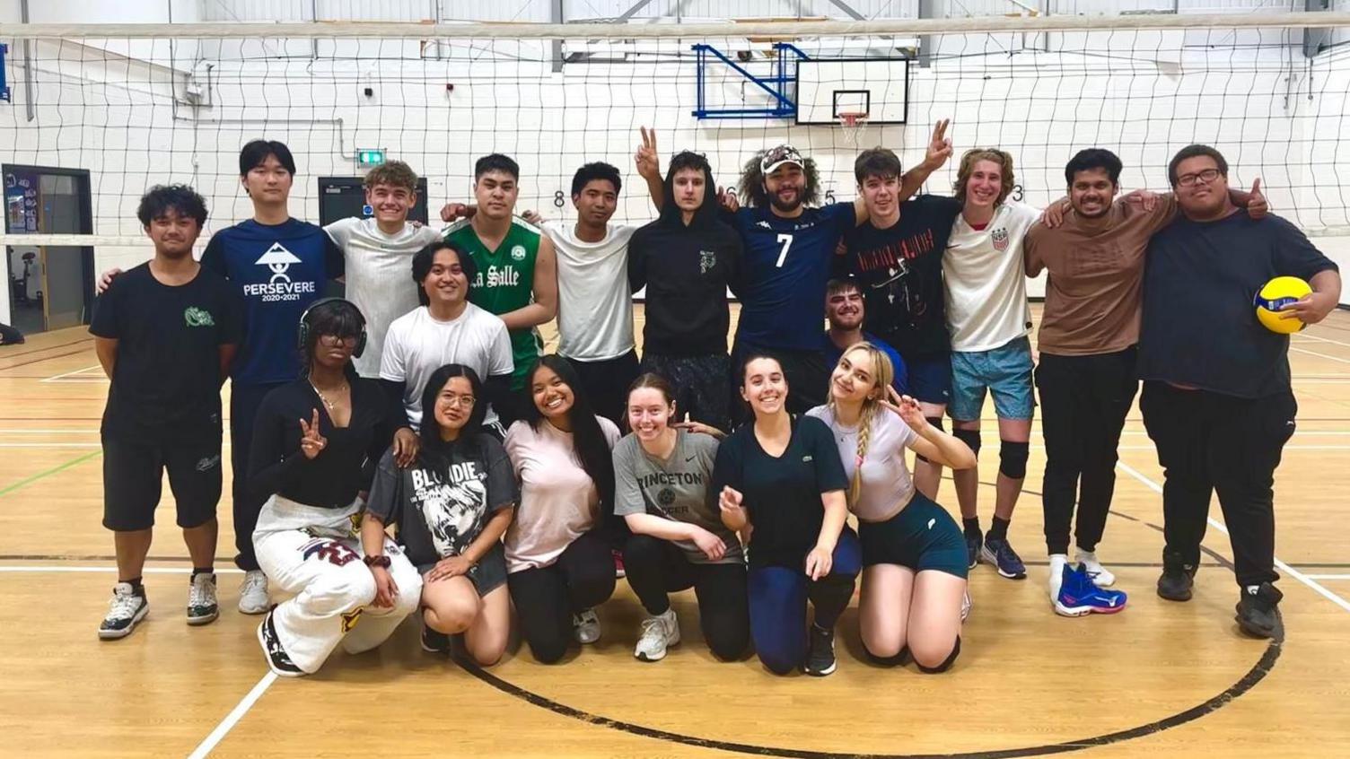 A group of students are standing in front of a volleyball net in a sports hall. They are all facing the camera.