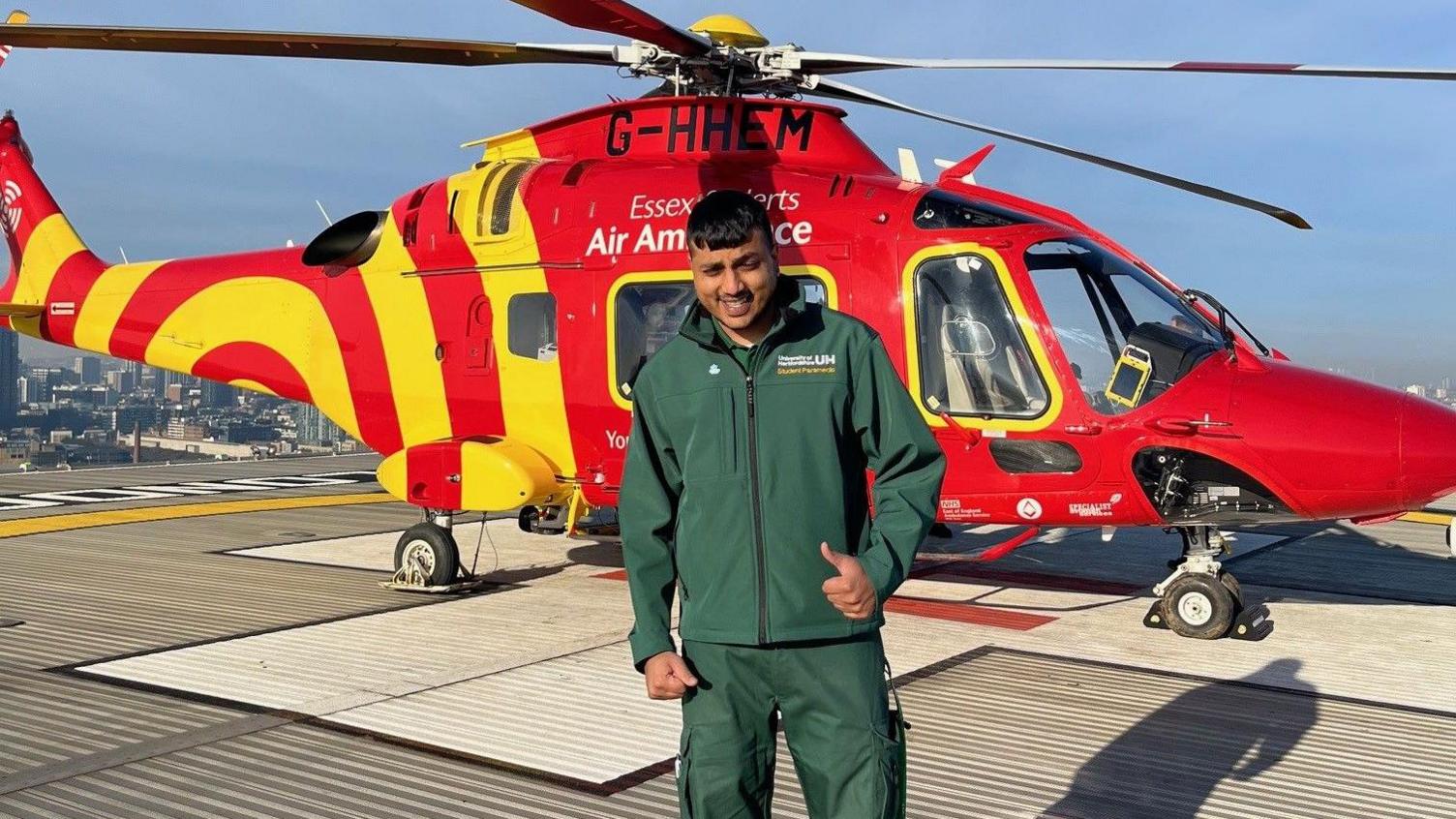 Jaabir Booly, wearing a green uniform, with top and bottoms, holding his left thumb up, standing in front of a red helicopter, with yellow stripes on it. 
