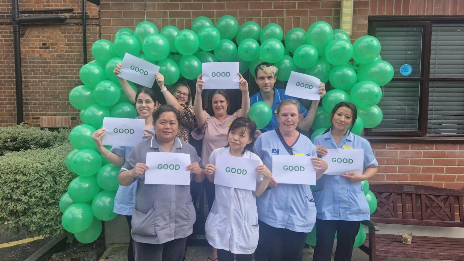  Eight members of staff are seen holding up Good signs in front of a set of green balloons. 