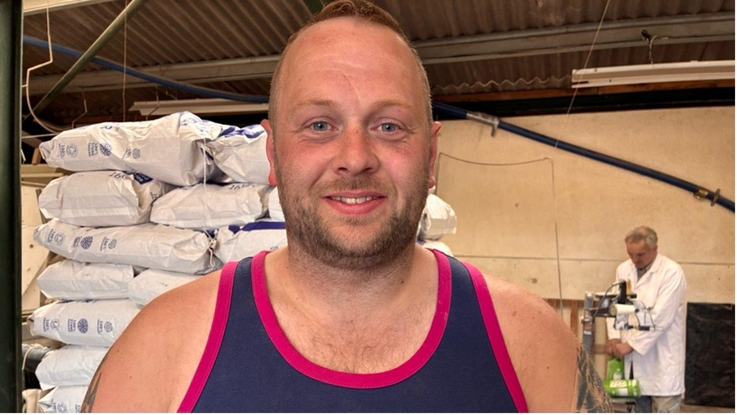 Robert Archer stands in the centre of the image wearing a blue vest with pink edging. Behind him is a stack of white bags of flour. To his right, a man in a white coat is working on a machine.
