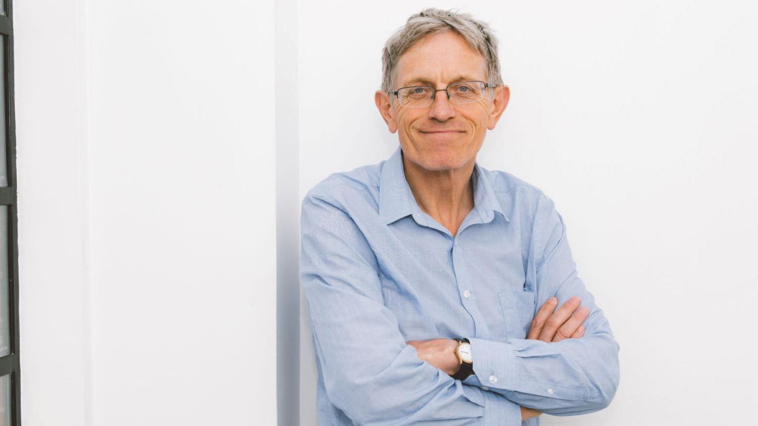 A smiling man with grey hair, wearing glasses, jeans and a blue, open-necked shirt leans against a white wall, with an old-industrial-style window to his right.