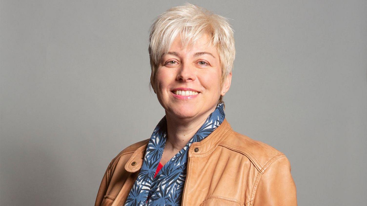 A woman with short blonde hair, a light brown jacket and a blue and white scarf, stands in front of a grey background and smiles.
