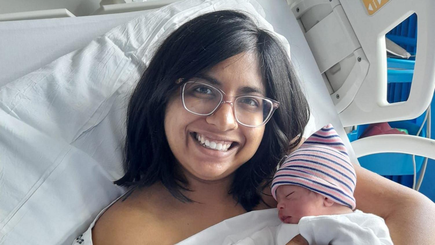 Priyanka Salamonczyk smiles at the camera while lying on a hospital bed and cradling a newborn. The baby is wearing a stripey hat and white baby grow. Priyanka has dark hair and is wearing glasses. 