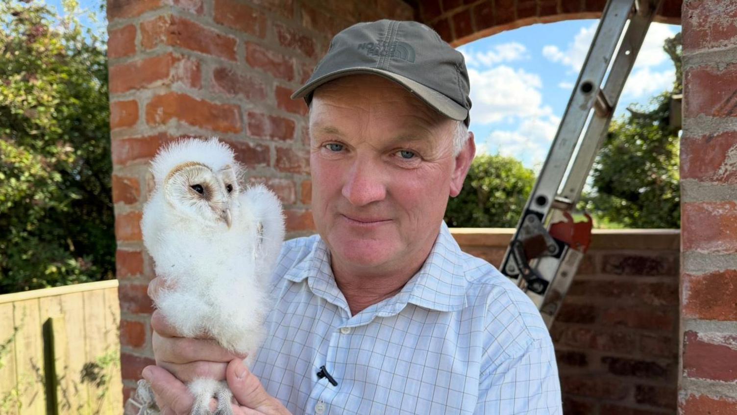 The photo shows a man looking at the camera. He's wearing a green baseball cap, and a white and blue checked shirt. To his left, he's holding a baby barn owl. It is covered in white, fluffy feathers and is looking to the side. He's standing in front of a brick building, which has a ladder resting against it.