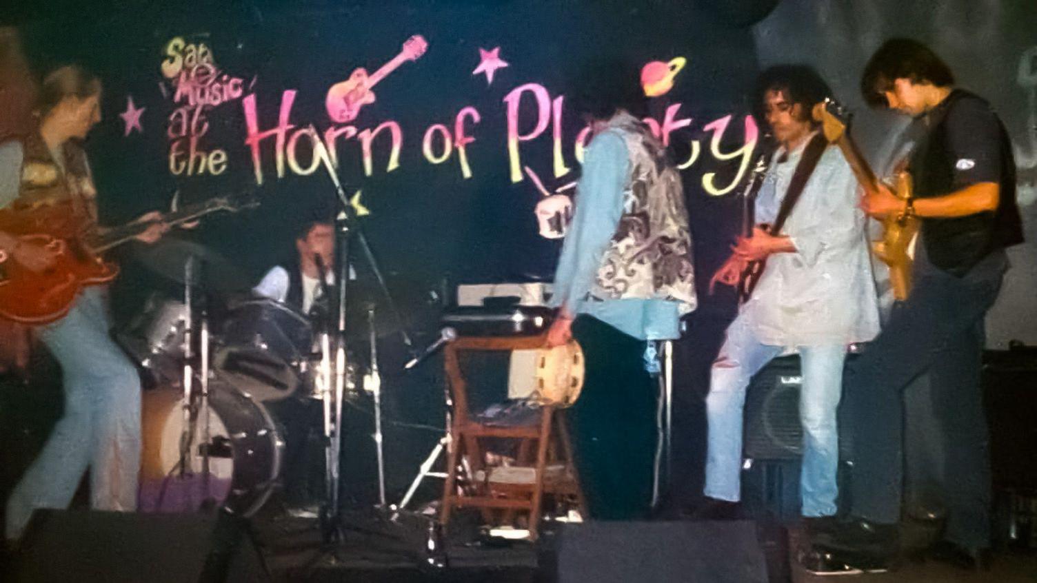 Five musicians on a stage in a slightly blurred image. Two men on the right are holding guitars, with a performer, with his back to the stage, holding a tambourine. A drummer is sitting down, and a guitarist is to the left.  A chair is in the middle and you can see a drum kit to the left. 