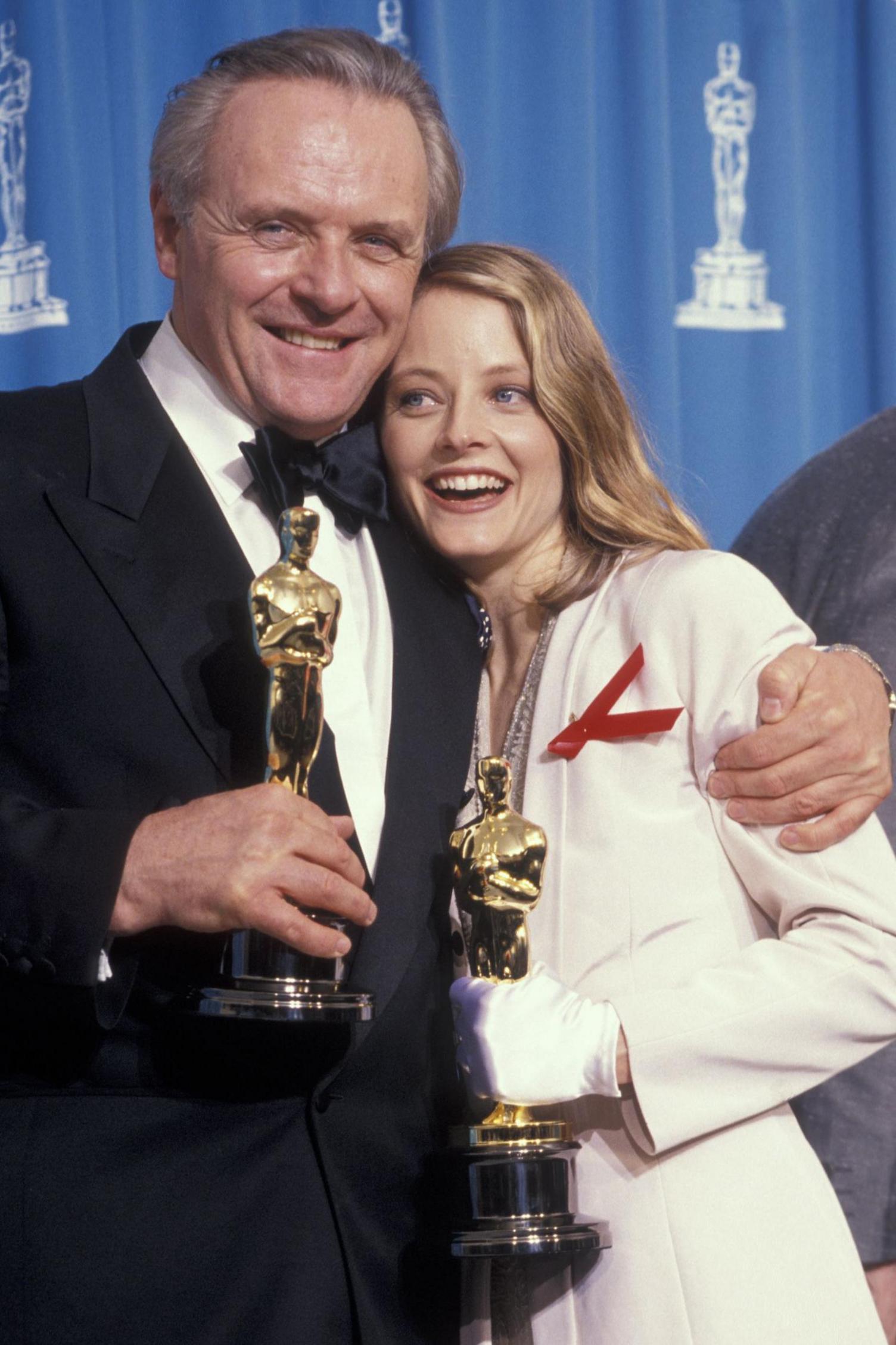 Hopkins in black dinner suit with his arm around Jodie Foster in white suit & gloves & red brooch both smiling and holding Oscars