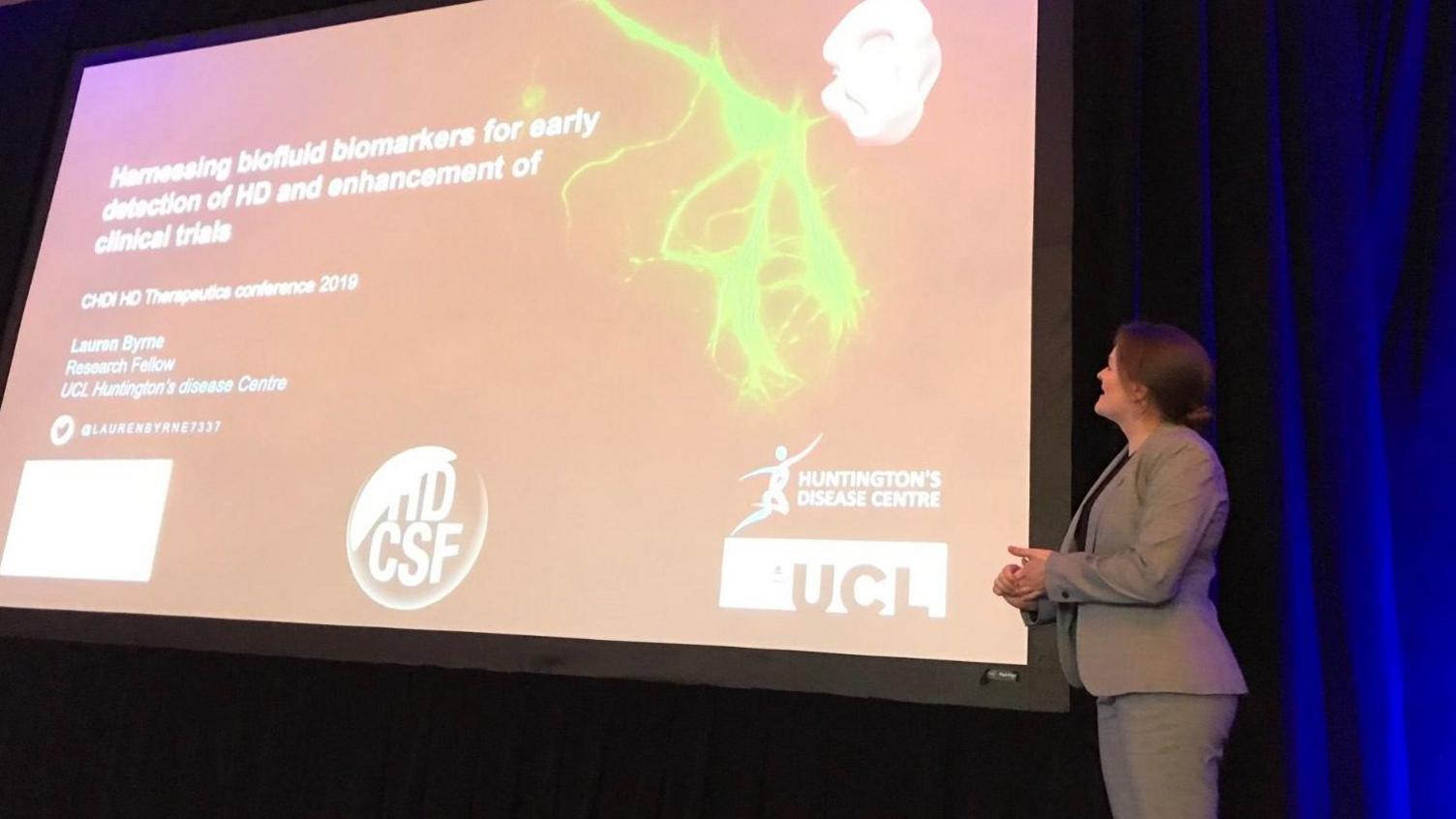Dr Lauren Byrne who has brown hair tied back and who is wearing a grey suit stands beside a large screen on a stage that contains information on a medical trial