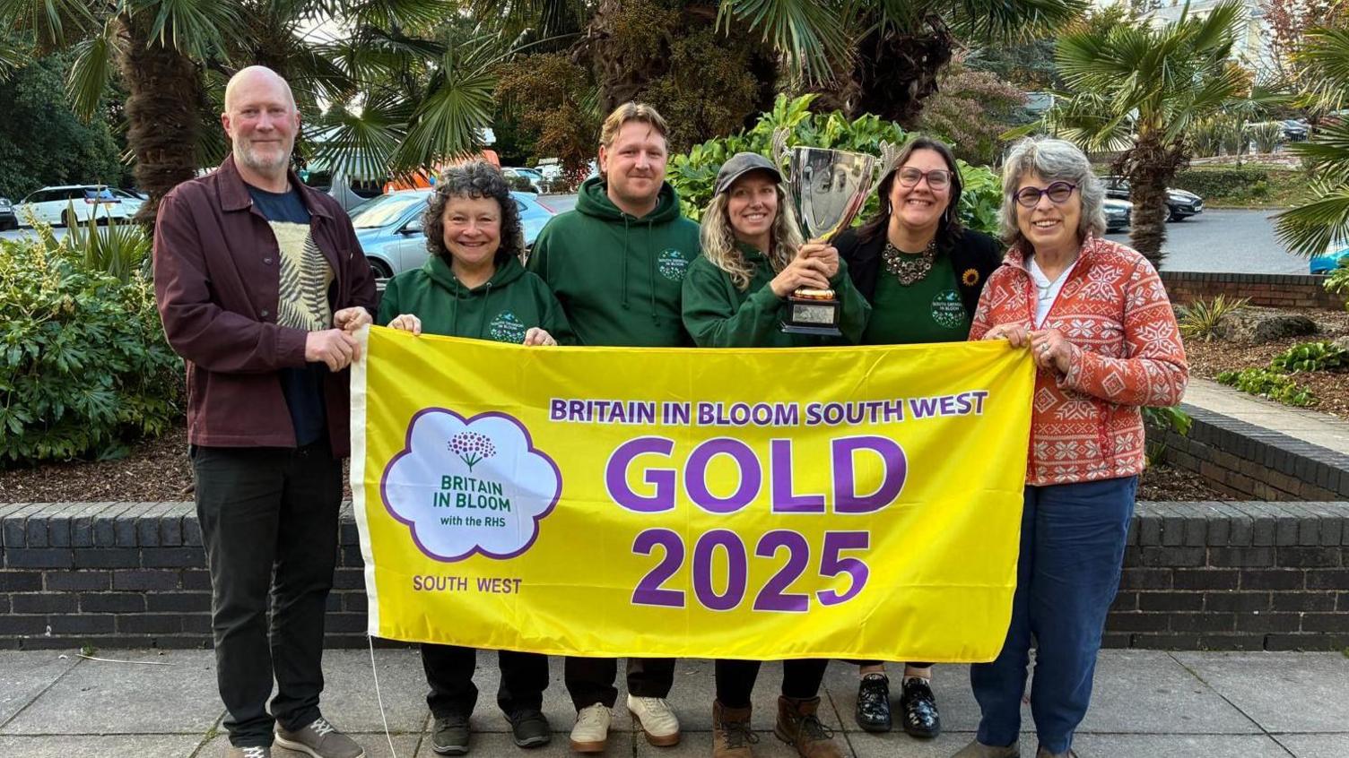 South Swindon in Bloom members smile by a car park behind a yellow flag which says 'Britain in Bloom South West Gold 2025'