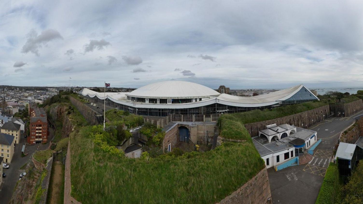 A wide shot of the Fort Regent leisure centre. It is a large white-roofed building surrounded by trees. There is a green hedges and grass around it along with solid stone walls and lots of houses. 