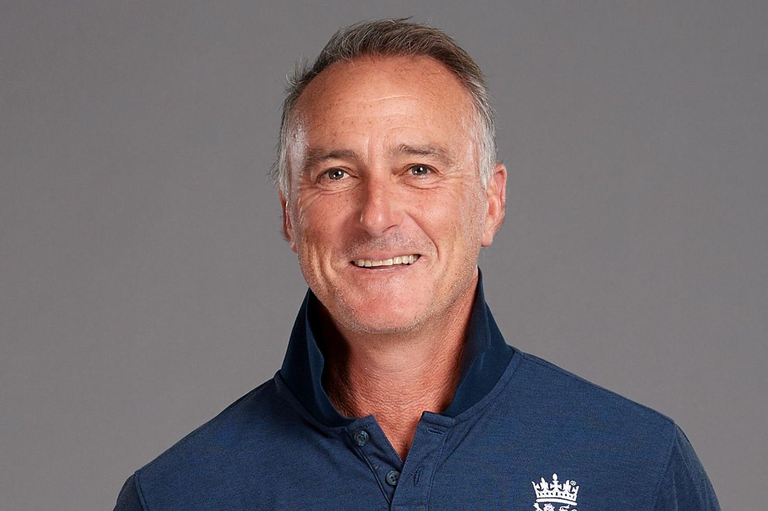 Graham Thorpe is pictured against a gray background - he has grey receding hair and a lively smile. He is wearing a dark blue long-sleeved polo top with what looks like the England logo . 