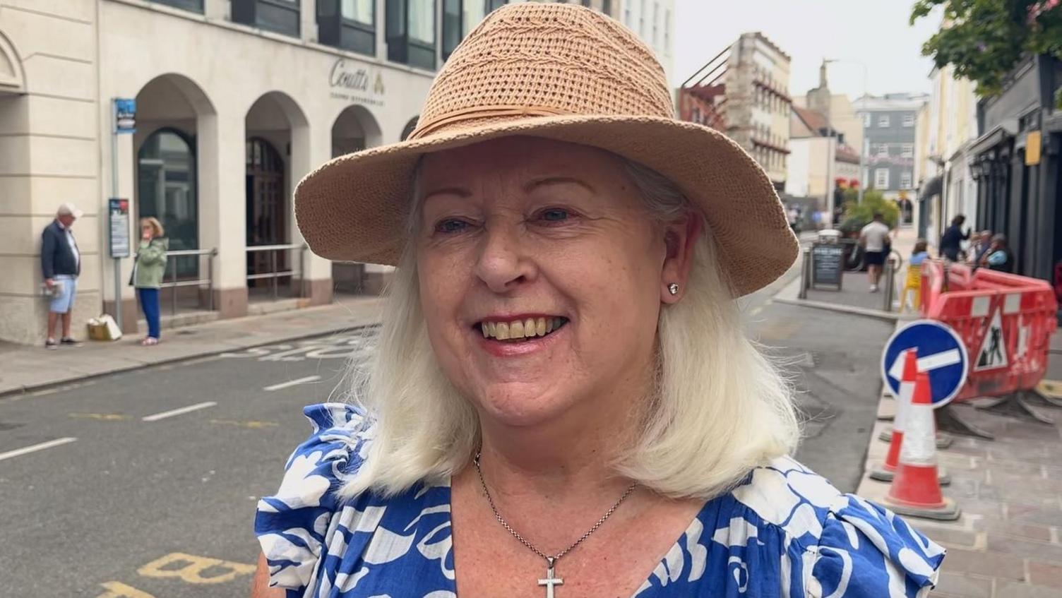 Jackie Hilton stands on Broad Street. Roadworks are visible behind her. She has shoulder length blonde hair and wears a blue dress and straw hat