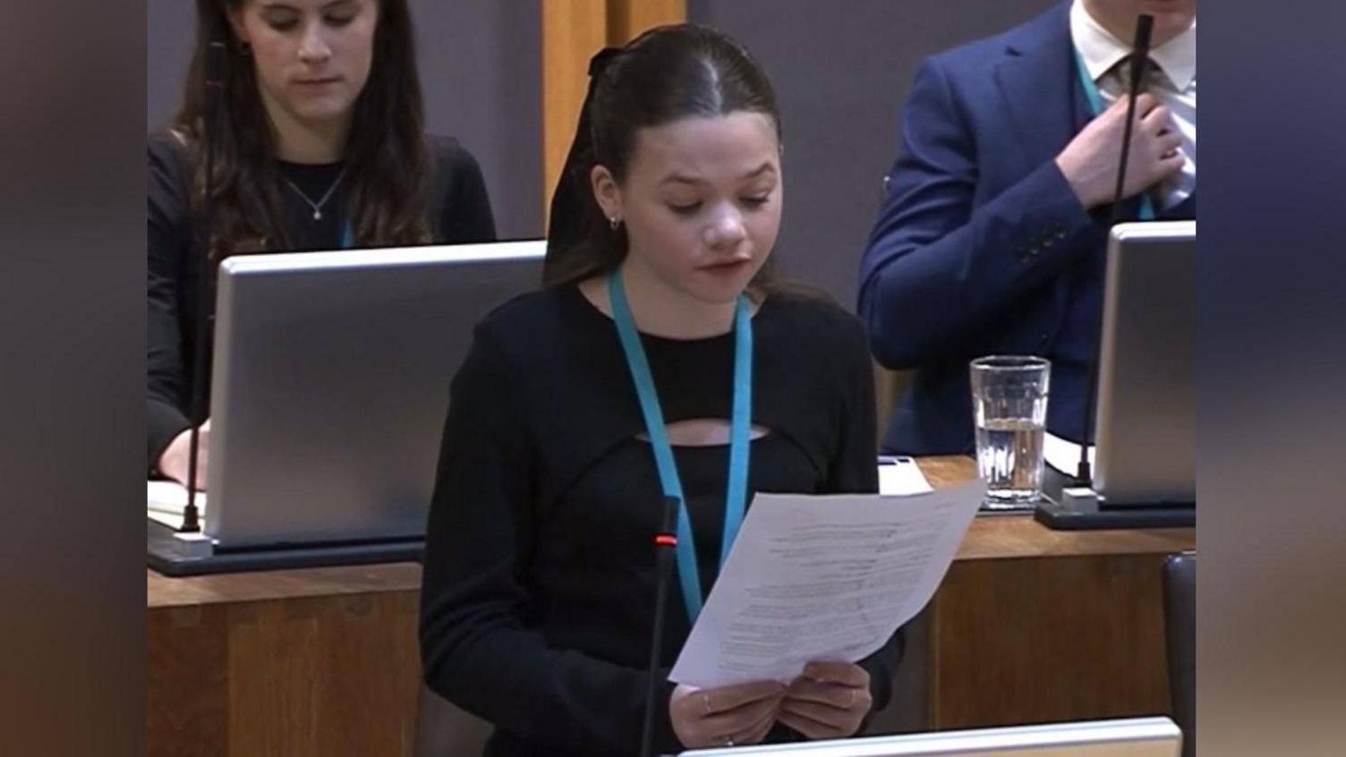 Llun o ferch yn ei harddegau yn sefyll yn darllen darn o bapur mae'n dal o'i blaen. Mae ganddi wallt brown. Mae'n gwisgo ffrog du. Mae yna ferch mewn siwmper du yn eistedd wrth ddesg tu ôl iddi yn edrych ar gyfrifiadur. Mae yna fachgen mewn siwt glas hefyd yn eistedd wrth ddesg tu ôl iddi.