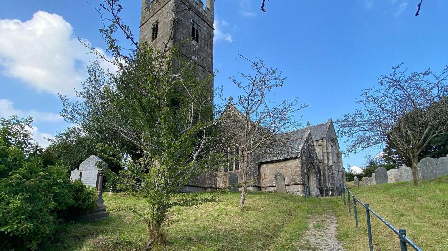 St Mary parish church's closed churchyard on a clear day. There are trees and large old gravestones. The grass is freshly mown and there are cuttings covering the path to the entrance. There is a metal handrail along the path.