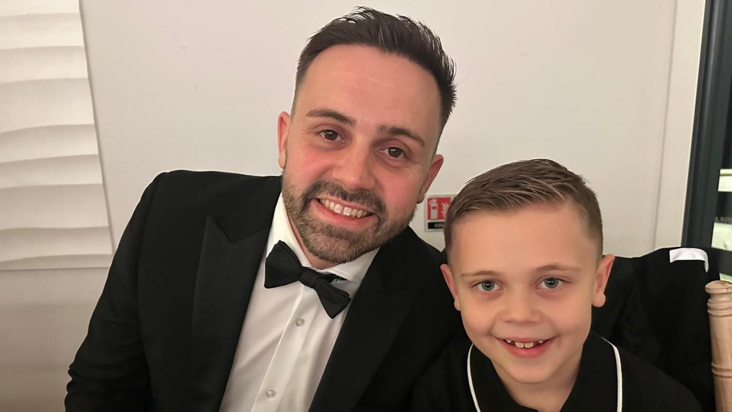 Neil Gilson, who is wearing a tuxedo with a black bow tie, smiles in a picture as he sits next to his son Jack, who is wearing a black polo shirt.