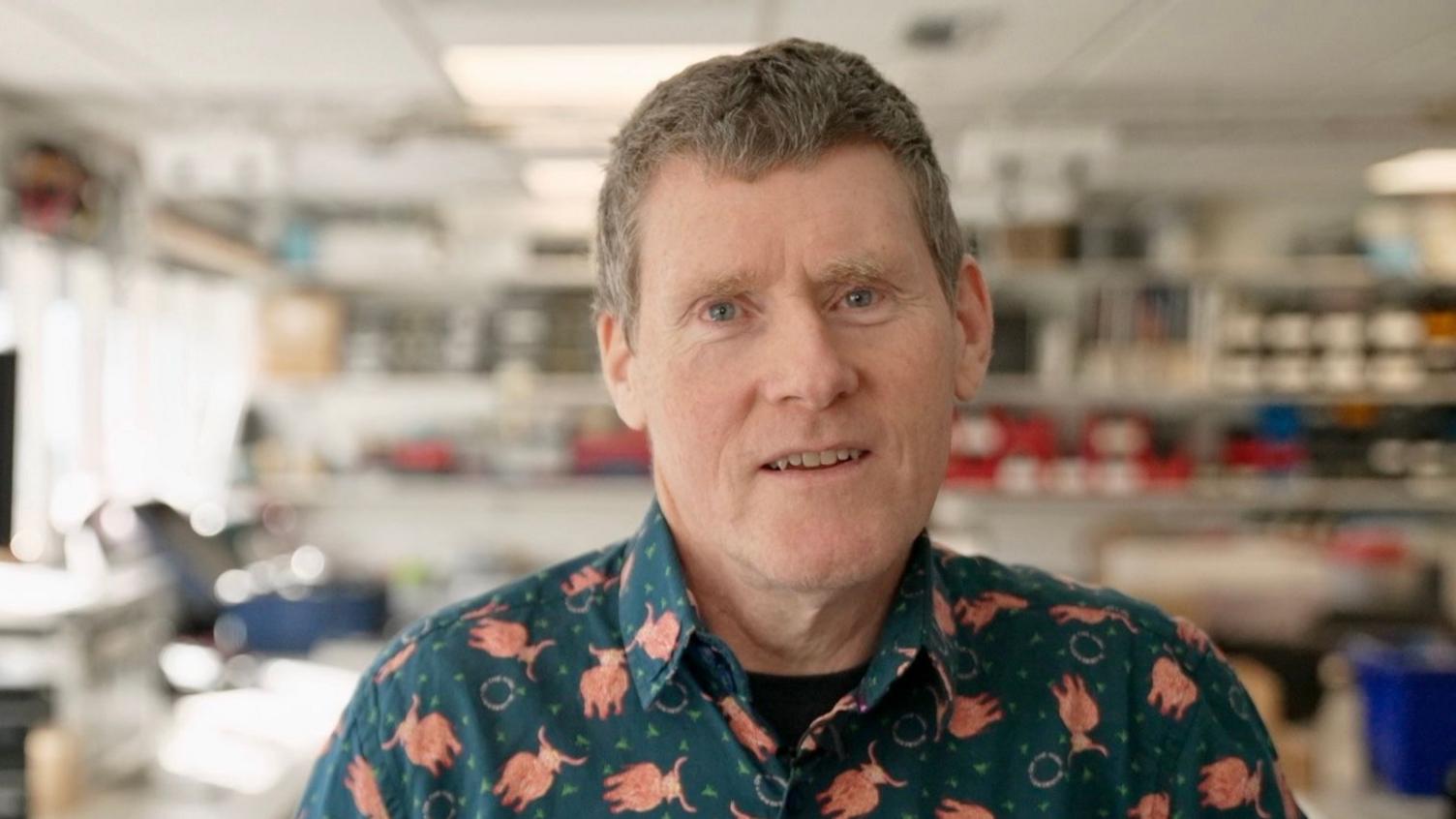 Dr Jim McQuaid is a middle aged man with short salt and pepper grey hair he stands in his lab at the university of Leeds wearing a green shirt with highland cows on it. Behind him are shelves of equipment but they are out of focus.
