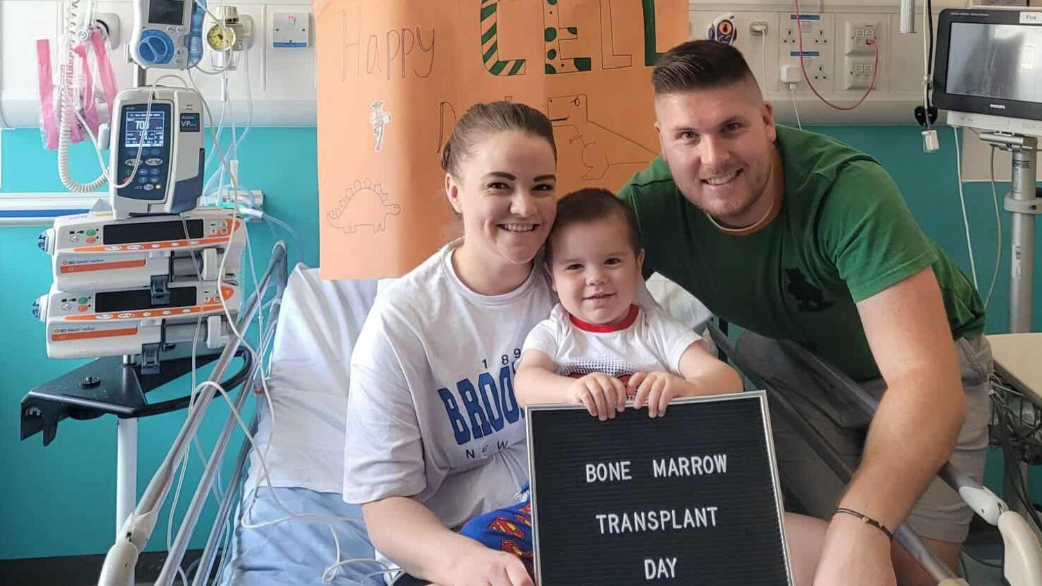 Louie, visibly younger in the photo, sits between his mum Shannon and dad Josh on a hospital bed, holding a board which reads 'bone marrow transplant day'. All three are smiling at the camera, and there is a poster behind them which says 'happy cell day' on orange paper. 