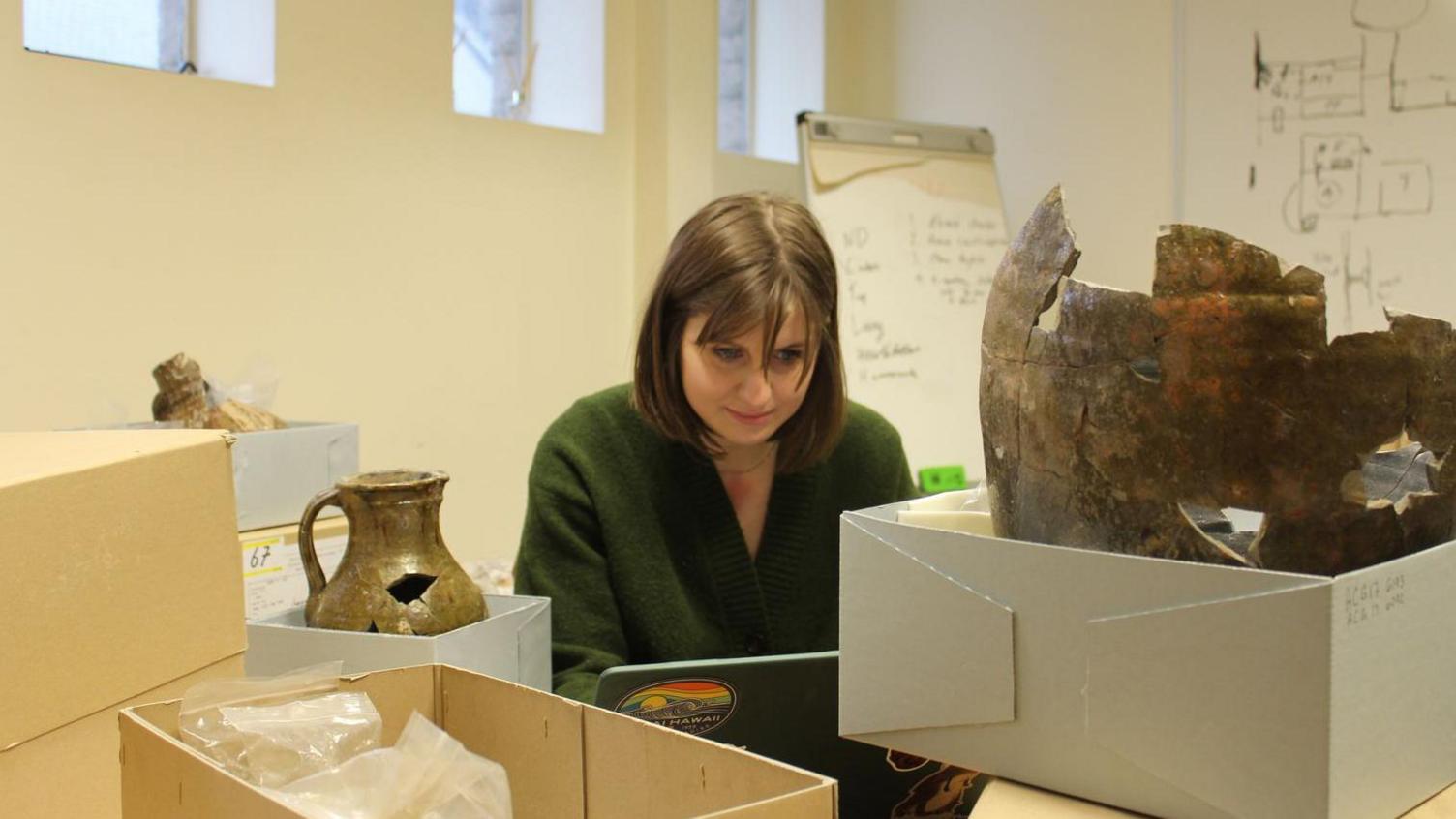 Doctoral Researcher Emily Walker typing on a laptop as she carries out some post-excavation work. She is surrounded by cardboard boxes. Some of the boxes are open, revealing metal and gold looking artifacts inside them. One of the items resembles a vase. Ms Walker is wearing a green cardigan. Her straight brown hair reaches her shoulders and she has a fringe. There is writing on the whiteboards behind her. 