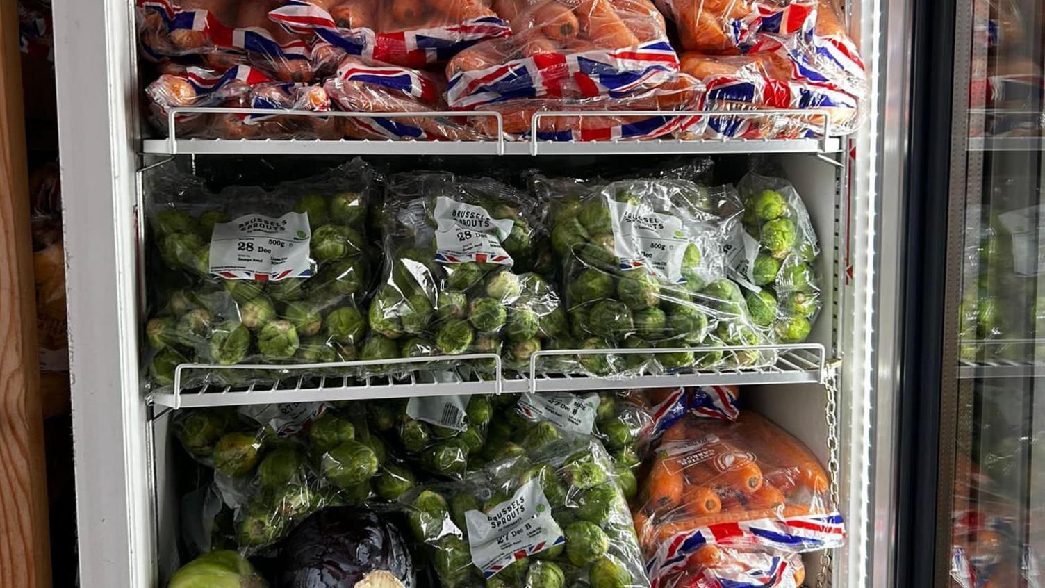 Three shelves of a large upright fridge full of bags of carrots and Brussels sprouts