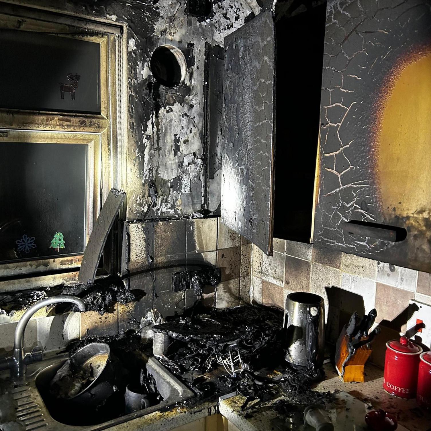 The corner of a kitchen after a fire. The cupboards are scorched black and the surfaces and sink are covered in ash.