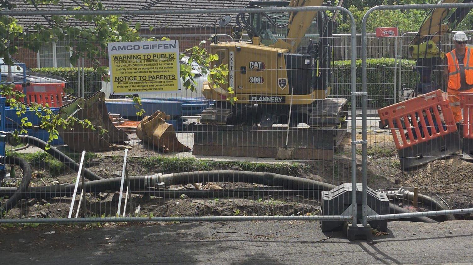 A view of construction site, behind a temporary wire fence, with black cables emerging from a hole in the road surface. A yellow digger is parked beyond the cables and a man in orange high vis uniform can be seen to the right. 