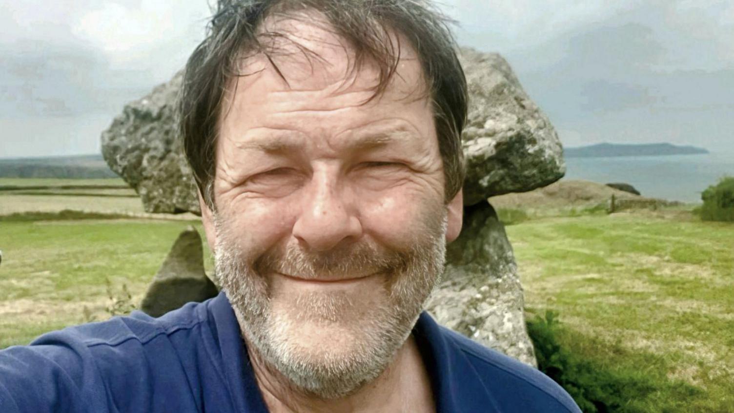 A smiling brown haired man with a white beard wearing a blue t-shirt, standing in front of a Neolithic tomb