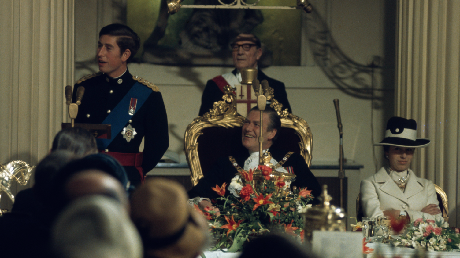 King Charles making a speech while standing next to the lord mayor of the City of London who is sitting in a grand seat. Princess Anne is wearing white and sitting next to him