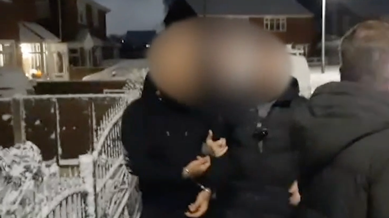 A man with his face blurred is handcuffed during an arrest in Wolverhampton. The arrest is taking place at night. The group of people is outside. They are all wearing black coats. There is ice and snow around them on railings and on shrubs.