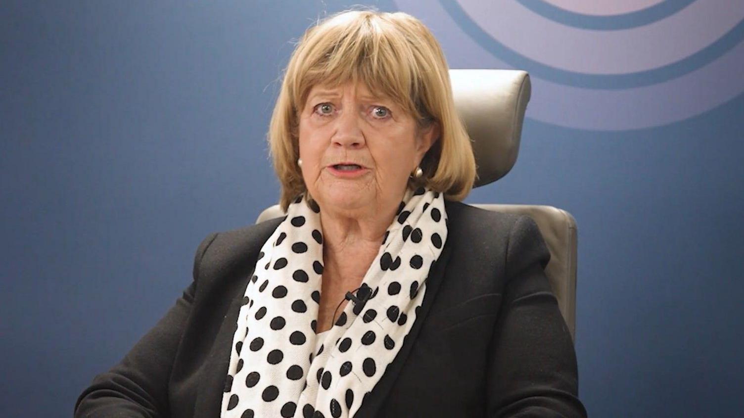 A woman with shoulder length blonde hair and a fringe. She is wearing a black blazer and a white and black polka dot scarf.