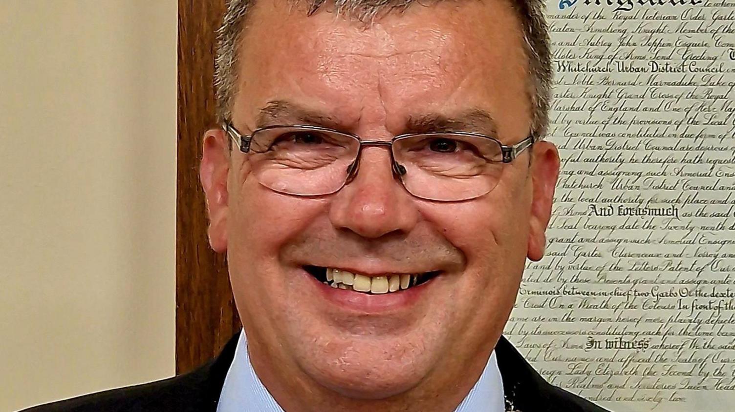 Close-up photo of a man smiling. He is wearing glasses, a shirt and suit jacket, and is in front of civic scripture framed on the wall behind him