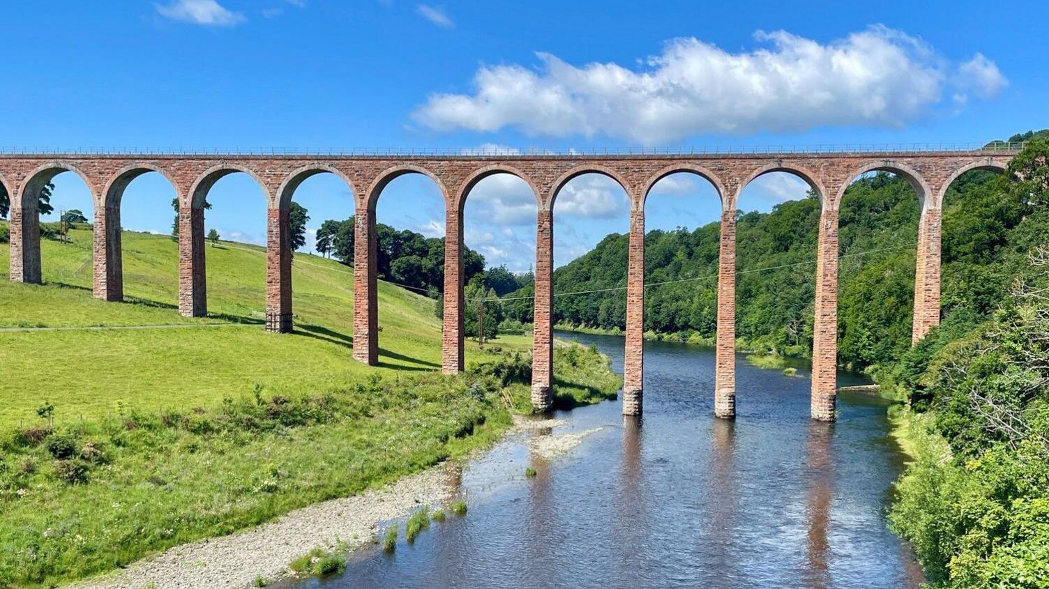 A viaduct with tall red-brick arches runs down a grassy hill and over a river and into a green forest on a bright and sunny day.